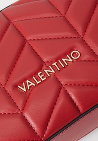 Red quilted leather handbag featuring a gold logo "VALENTINO" and contrasting black trim. The surface has a smooth texture with stitched detailing.