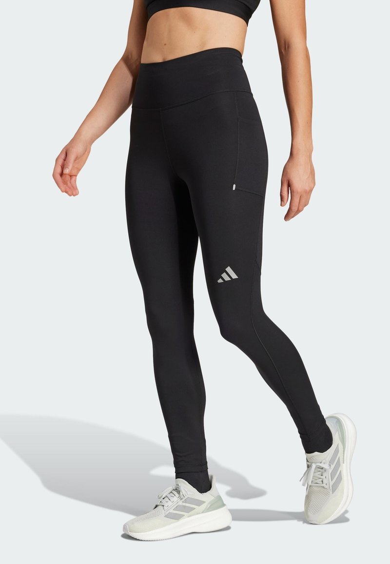 Zwarte high-waisted leggings van rekbaar materiaal, met een zijzak en een grijs logo op het linkerpijpje, gedragen met lichte sportschoenen.