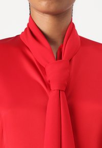 Rød bluse med en glatt satinfinish, med en knyttet halsdetalj som faller elegant. Stoffet er lett med en rik farge.