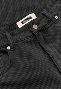 Gros plan sur la ceinture texturée noire d'un jean avec bouton en métal et rivets, et une étiquette blanche affichant le nom de la marque "WOODBIRD."