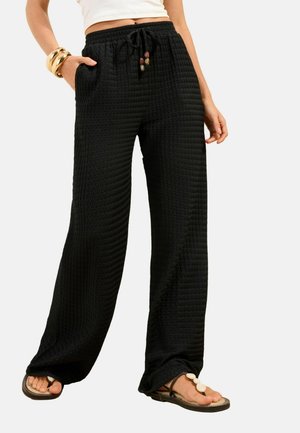 Kobieta ubrana w czarne, teksturowane spodnie o szerokich nogawkach ze sznurkiem i koralikami, biały krótki top, złote bransoletki oraz brązowe sandały z dekoracją z muszelek.