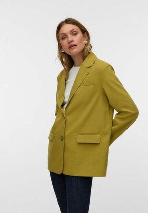Kvinde iført oversized olivengrøn blazer, hvid top, mørke jeans og sølvøreringe, stående med den ene hånd bag ryggen mod hvid baggrund.