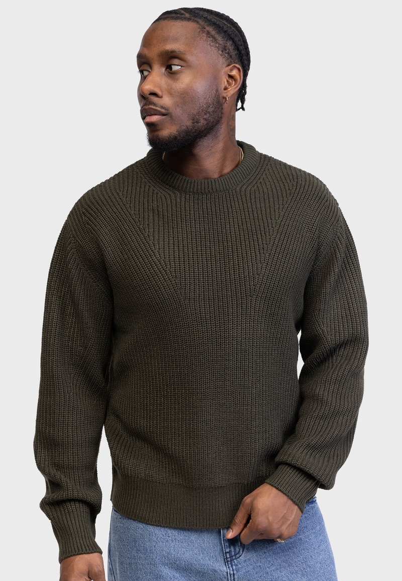 Suéter de punto verde oscuro con un patrón texturizado, cuello redondo y puños de canalé. El tejido parece grueso y cálido. Combinado con jeans azul claro.