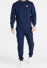 Mörkblå sweatshirt och matchande joggers i mjukt tyg. Har ribbade muddar och en liten logotyp på bröstet. Vita sneakers fulländar looken.