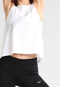 Mujer levantando los brazos, vestida con una camiseta sin mangas blanca de Nike y leggings negros de Nike, frente a un fondo liso.