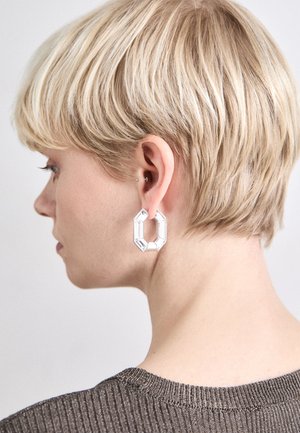 Silver geometric earrings, made of transparent acrylic, with a hexagonal shape and polished finish, worn on short hair.