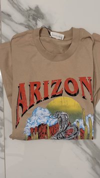 Bežs kokvilnas T-krekls ar uzrakstu "ARIZONA" novecotā sarkanā krāsā, zem kura ir krāsains attēls ar čūsku un tuksneša ainavu.