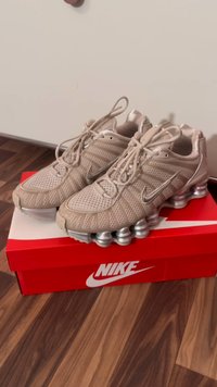 Pantofi sport Nike bej cu textură din plasă și accente argintii pe talpă, așezați pe o cutie roșie Nike pe un podea din lemn.