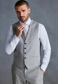 Gilet gris ajusté avec six boutons noirs, porté par-dessus une chemise blanche et une cravate à motifs, avec un tissu lisse et une coupe structurée.