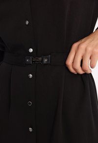 Czarna sukienka wykonana z fakturowanej tkaniny, z zapięciem na guziki z przodu oraz paskiem z metalowym zapięciem. Projekt zawiera akcenty w postaci srebrnych guzików.