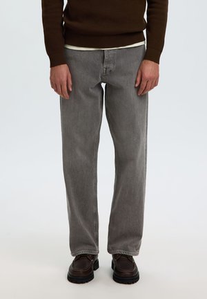 Mann trägt graue Straight-Leg-Jeans, braune Loafers und einen braunen Pullover mit sichtbarem weißen Hemdsaum, steht vor einem schlichten Hintergrund.