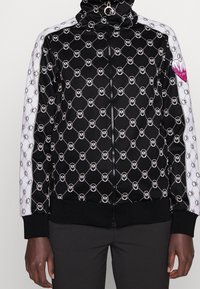 Black zip-up jacket with all-over circular pattern, white side panels, high collar, and pink heart accent on the chest. Textured fabric.