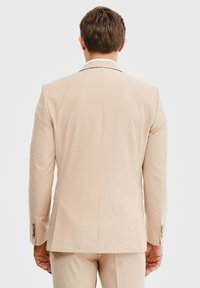 Jasno-beżowa marynarka w fasonie slim fit z teksturowanym wykończeniem, z klapami i guzikami na rękawach. Widok z tyłu.