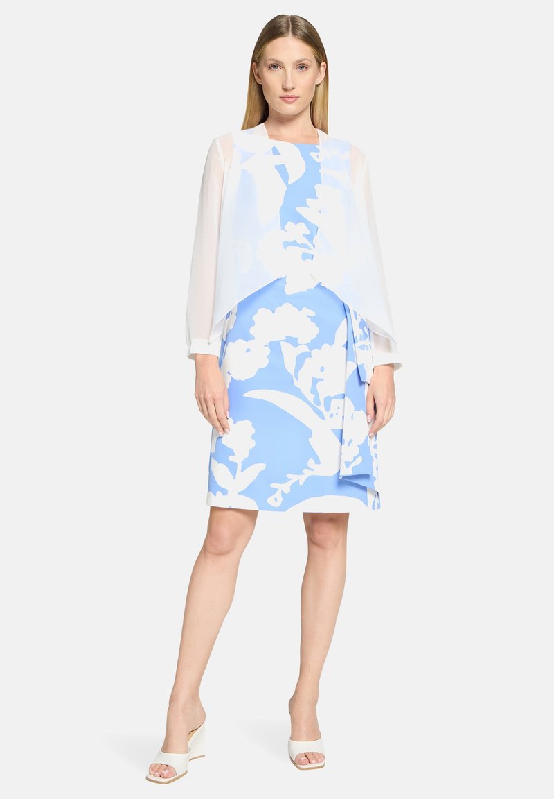 Une superposition légère blanche transparente à manches longues sur une robe bleue ornée de grands motifs floraux blancs, complétée par des sandales blanches à talons.