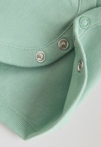 Krupan plan svijetlo zelene tkanine sa srebrnim drukerima, prikazujući šavove i rebraste rubove na otvaranju odjeće.