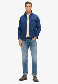 Giacca di jeans blu con colletto alto, chiusura con bottoni e una tasca sul petto, abbinata a jeans azzurri e stivali marroni con lacci.