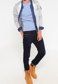 Grijze rits hoodie over een blauwe t-shirt, gecombineerd met donkere jeans en beige veterschoenen. Kenmerken zijn onder andere contrasterende denim accenten en opgerolde mouwen.