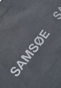 Primer plano de una tela gris oscura con el texto blanco "SAMSØE" impreso, mostrando la textura de la tela y los detalles de la costura.