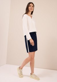 Kvinna som går klädd i en vit långärmad tröja, marinblå kjol med sidostrip, och beige sneakers mot en enkel, ljus bakgrund.