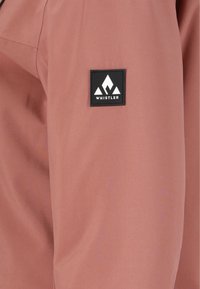 Veste rose clair avec une texture de tissu, un patch noir de marque avec un logo de montagne blanc sur la manche gauche.