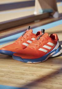 Pomarańczowe buty sportowe z białymi paskami, siatkową cholewką, teksturowanym wykończeniem oraz niebieskimi akcentami na podeszwie i pięcie. Posiadają pętelkę do zakładania.