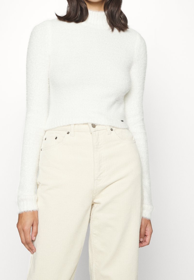 Femme portant un pull court à manches longues blanc et duveteux ainsi qu'un pantalon en velours côtelé crème taille haute, debout devant un fond blanc.