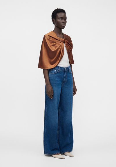 Femme portant un capelet en satin couleur rouille, un haut blanc, un jean bleu taille haute à jambes larges et des chaussures blanches pointues, debout sur un fond uni.