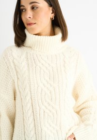 Maglione in crema con lavorazione a maglia grossa e colletto a costine alto, caratterizzato da motivi a treccia e maniche testurizzate. Tessuto morbido e caldo.
