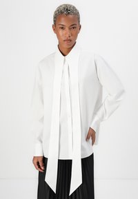 White long-sleeve shirt with a structured collar and a full-length tie detail. Smooth texture, button-down front, paired with a pleated black skirt.
