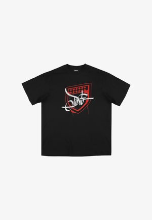 Czarna koszulka z bawełny o luźnym kroju, z czerwoną grafiką w kształcie tarczy i białym, stylizowanym tekstem, akcentowana szczegółami w postaci rozpryskanego farby.