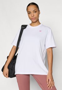 Bijela majica opuštenog kroja s malim ružičastim logotipom. Nošena s lagano ružičastim tajicama i crnom torbom preko ramena. Glatka tekstura.