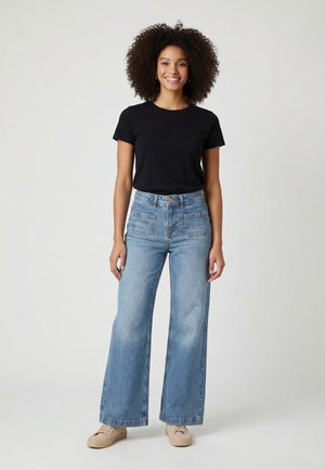 Mujer sonriente con cabello rizado, vistiendo una camiseta negra, vaqueros azules de tiro alto y pierna ancha, y zapatillas beige, de pie frente a un fondo liso.