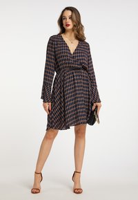 Rochie înfășurată de culoare albastru închis, cu mâneci lungi, model floral în tonuri de aur și rugină, talie ajustată, fustă evazată, asortată cu o poșetă neagră și pantofi cu toc maron.