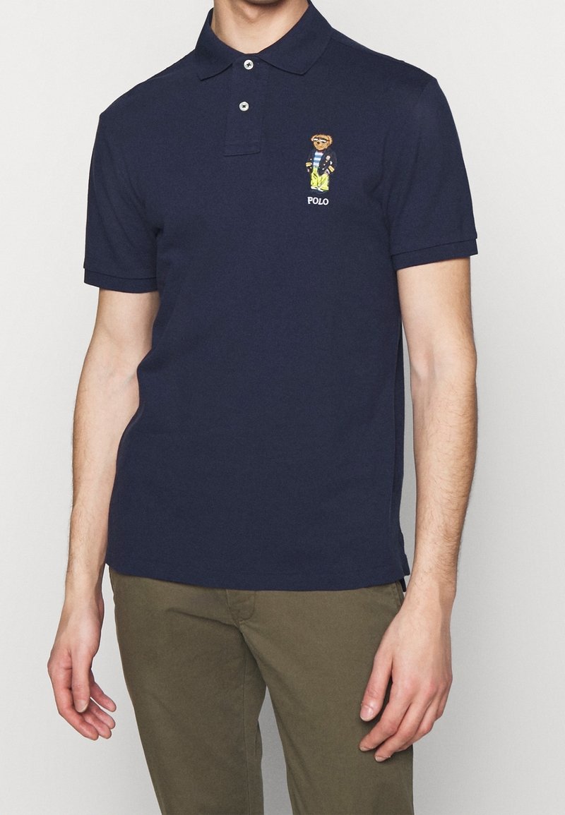 Homme portant un polo bleu marine avec un petit ours brodé en veste et pantalon jaune sur la poitrine, assorti à un pantalon vert olive.