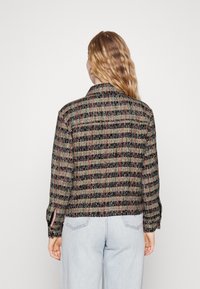 Woman with wavy red hair wearing a black, green, and red plaid jacket and light blue jeans, shown from the back on a white background.