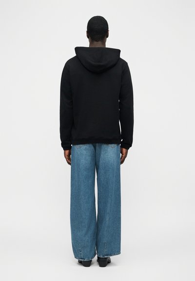 Άνθρωπος που στέκεται με την πλάτη γυρισμένη, φοράει μαύρο φούτερ με κουκούλα και φαρδιά μπλε τζιν παντελόνια, σε απλό ανοιχτό γκρι φόντο.