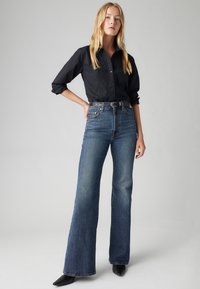 Black button-up shirt with floral embroidery, high-waisted, flared blue jeans, and pointed black heels. Smooth denim texture, tailored fit.