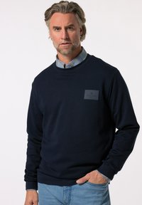 Sweatshirt bleu marine à col rond, avec un tissu texturé, des poignets élastiques et un patch logo sur la poitrine. Associé à une chemise à carreaux.