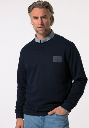 Felpa blu navy con collo a giro, realizzata in tessuto texturizzato, con polsini elasticizzati e una toppa con logo sul petto. Abbinata a una camicia a quadri.