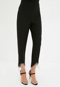 Czarne spodni z tapered fit i asymetrycznymi dołami ozdobione wiszącą frędzlą z kryształków, w połączeniu z czarnymi sandałami na wysokim obcasie z paskami wysadzanymi kryształkami.