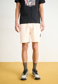 Beige shorts with a side pocket and a black label, paired with a black graphic t-shirt, gray socks, and multicolored sneakers.