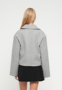 Γυναίκα με μακριά ξανθά μαλλιά που φοράει γκρι μάλλινο σακάκι και κοντή μαύρη φούστα, γυρισμένη με την πλάτη σε λευκό φόντο.