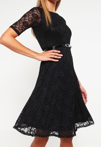 Musta pitsimekko, jossa on lyhyet hihat, istuva yläosa ja levenevä hame. Sisältää kiiltävän mustan vyön vyötäröllä ja kukkakuvioista pitsiä.