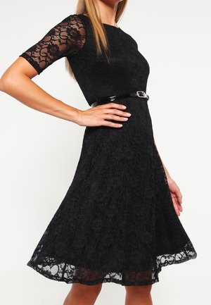 Abito in pizzo nero con maniche corte, corpetto aderente e gonna svasata. Presenta una cintura nera lucida in vita e un motivo floreale in pizzo.