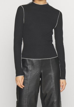 Persoon die een zwart shirt met lange mouwen draagt met witte naaddetails en zwarte leren broek met verticale stiknaden.