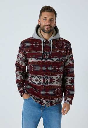 Jachetă cu model roșu și gri, cu glugă, având nasturi și un material texturat. Designuri geometrice și tribale notabile pe întreaga suprafață.