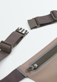Sangle élastique marron avec boucle à libération latérale en plastique et section en tissu beige avec poche zippée sur fond blanc.
