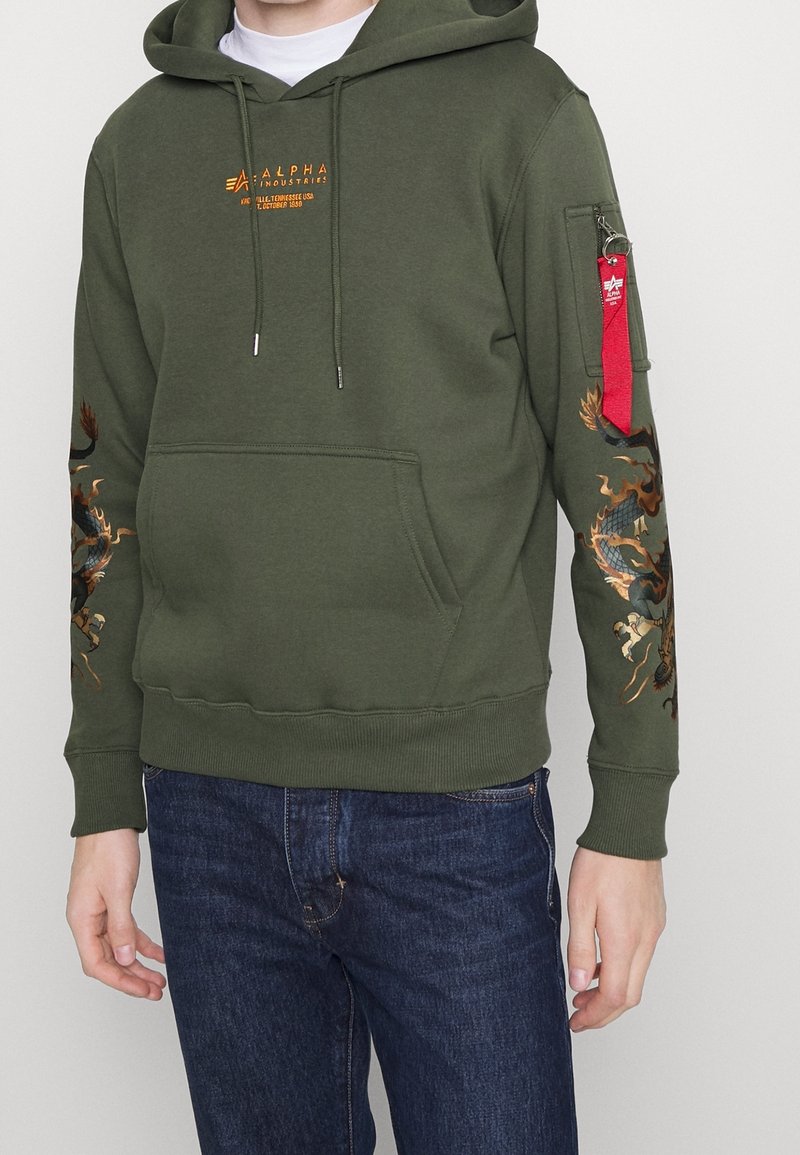 Sudadera con capucha de color verde oliva que presenta un bolsillo tipo canguro en el frontal, un logo bordado y un gráfico de dragón en la manga. Bolsillo con cremallera con acento rojo.
