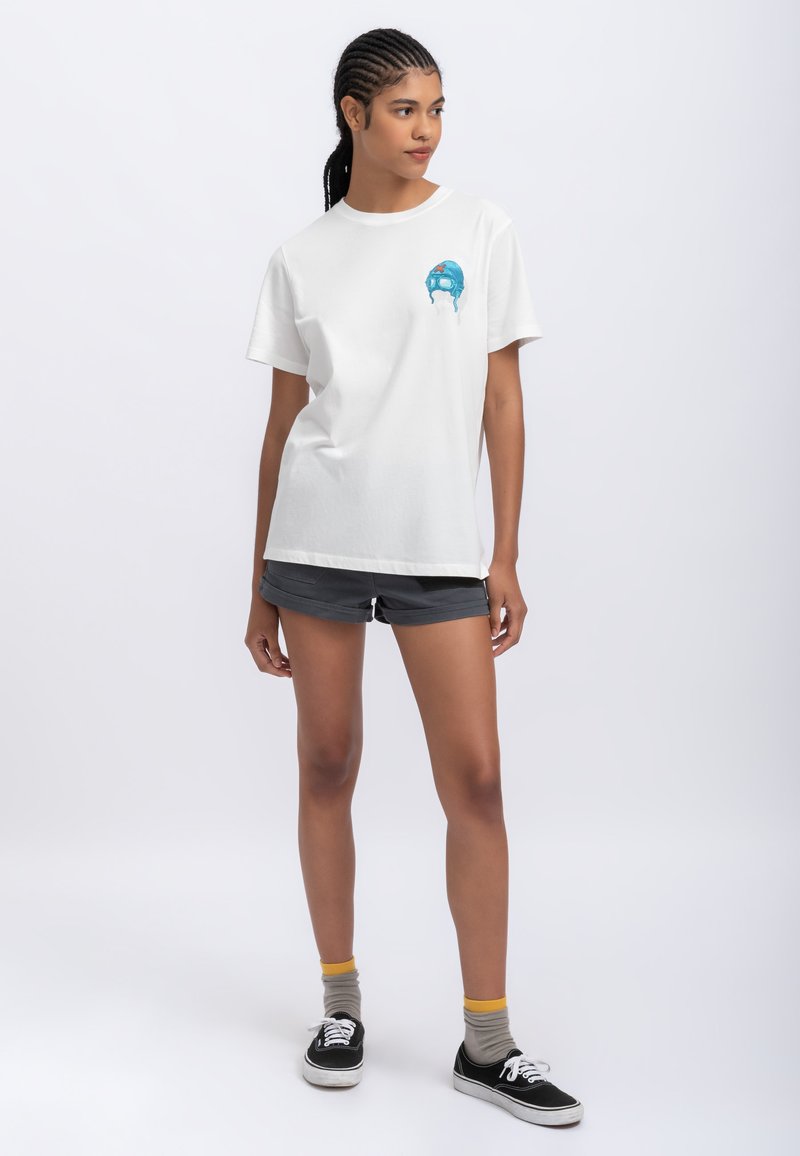 T-shirt bianca in cotone con un design grafico blu sul petto. Abbinata a pantaloni shorts grigi e sneakers nere. Sfondo semplice.