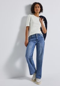 Haut blanc à manches courtes associé à un jean large bleu. Le modèle porte des baskets blanches et grises, se tenant devant un fond neutre.